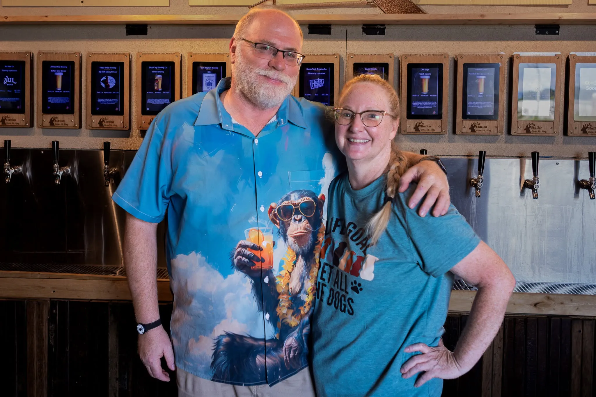 Lori and Brian standing arm in arm in front of the self-pour tap wall at Echo Station