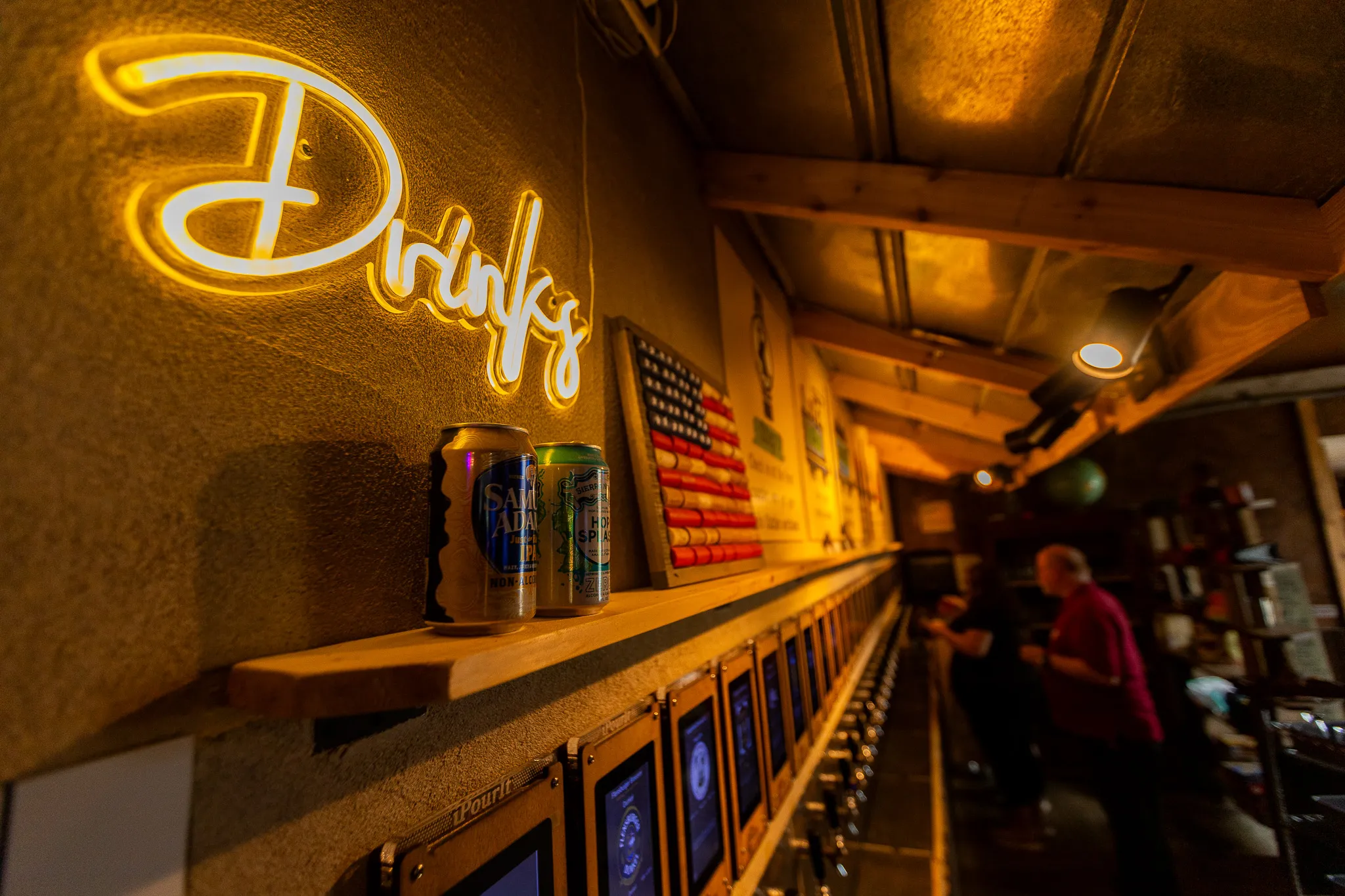 The self-pour tap wall, lit by a neon "Drinks" sign, with wood-framed screens above each tap